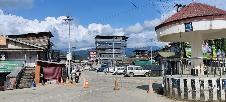 A street in Itanagar town with shops, parked cars and green hills under white clouds