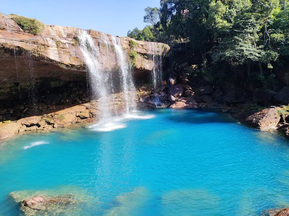 Bright turquoise pool at the foot of Krang Suri Falls framed by red rock and trees