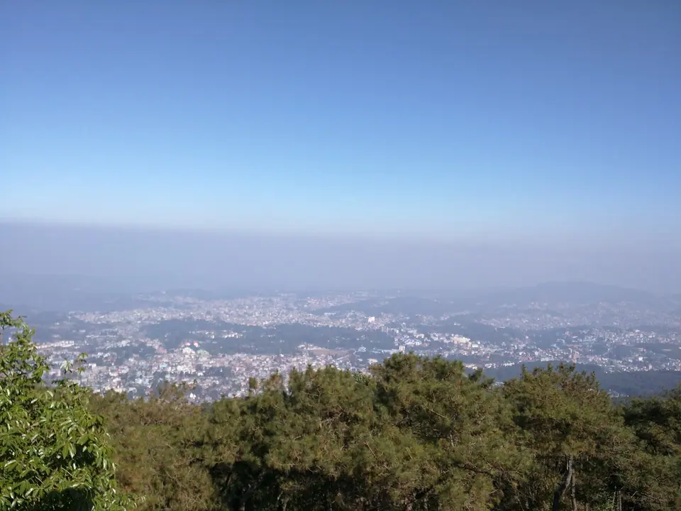 Shillong city in blue haze below a forested slope, seen from Shillong Peak on a clear morning