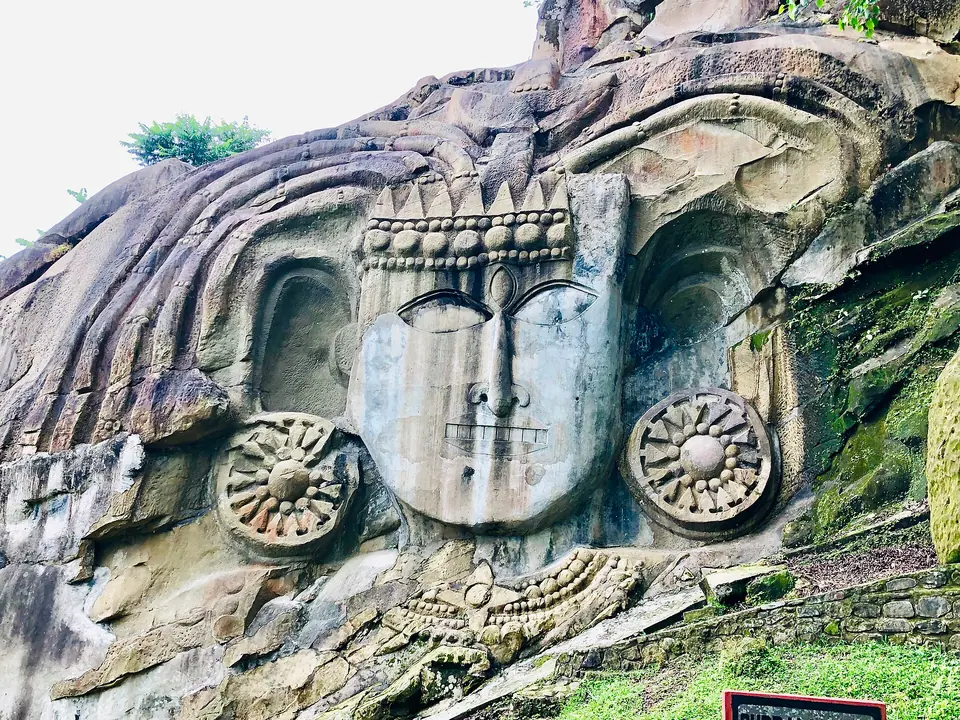 A crowned stone head with large disc earrings carved into the rock wall at Unakoti