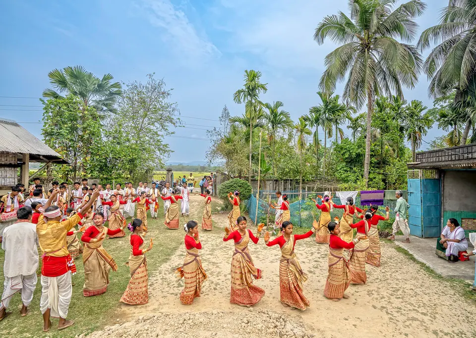 Women in red and cream mekhela sador dancing Bihu in a village field fringed with palms, Assam