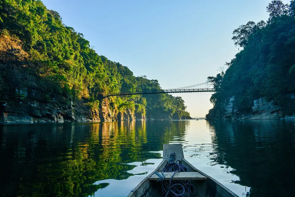 A wooden boat on the glassy Umngot River at Dawki, between forested cliffs and a suspension bridge